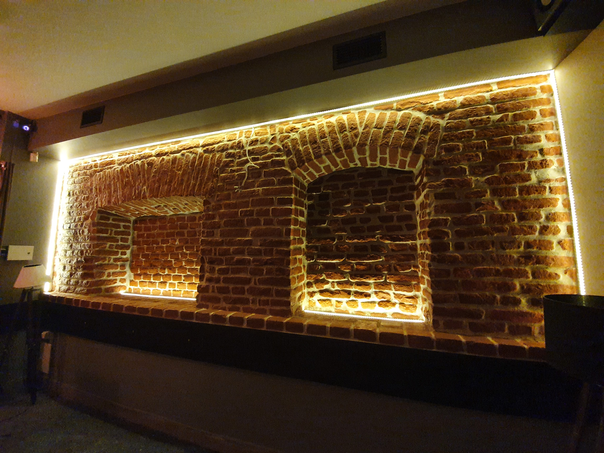 Ceglana ściana z dwoma wnękami, podświetlona ledami, tworząca unikalny element dekoracyjny wnętrza. Widoczne nierówności i faktura cegieł.