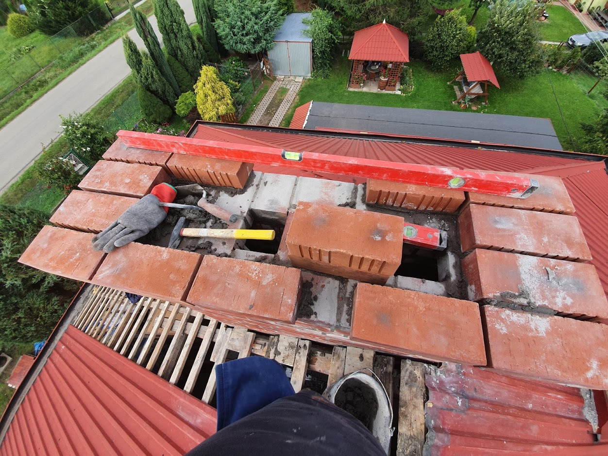 Murowanie komina z czerwonej cegły na dachu z blachy, widok z góry. Poziomica, kielnia, młotek i rękawica robocza obok. Prace murarskie w toku.