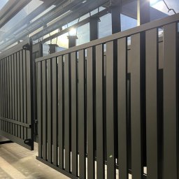 Malar - Szare, pionowe elementy ogrodzenia metalowego w hali produkcyjnej, zawieszone na szynach. Widoczne refleksy światła na powierzchni, podkreślające gładką fakturę.