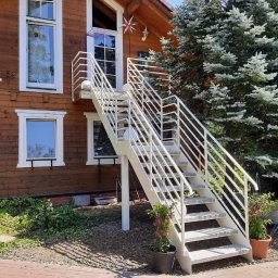 Malar - Białe schody zewnętrzne z metalową balustradą prowadzące do drzwi na piętrze drewnianego domu, otoczone zielenią i kwiatami w donicach. Ujęcie z dołu.