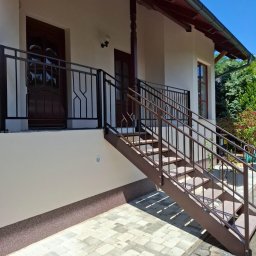 Malar - Zewnętrzne schody metalowe z balustradą o geometrycznym wzorze, prowadzące do drzwi wejściowych domu. Brązowa konstrukcja na tle jasnej elewacji i kostki brukowej.