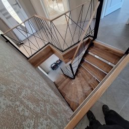 Malar - Spiralne schody z drewnianymi stopniami i nowoczesną balustradą z czarnych, krzyżujących się prętów, widziane z góry, z fragmentem osoby w kadrze.