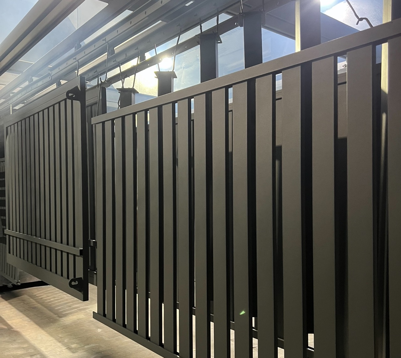 Szare, pionowe elementy ogrodzenia metalowego w hali produkcyjnej, zawieszone na szynach. Widoczne refleksy światła na powierzchni, podkreślające gładką fakturę.