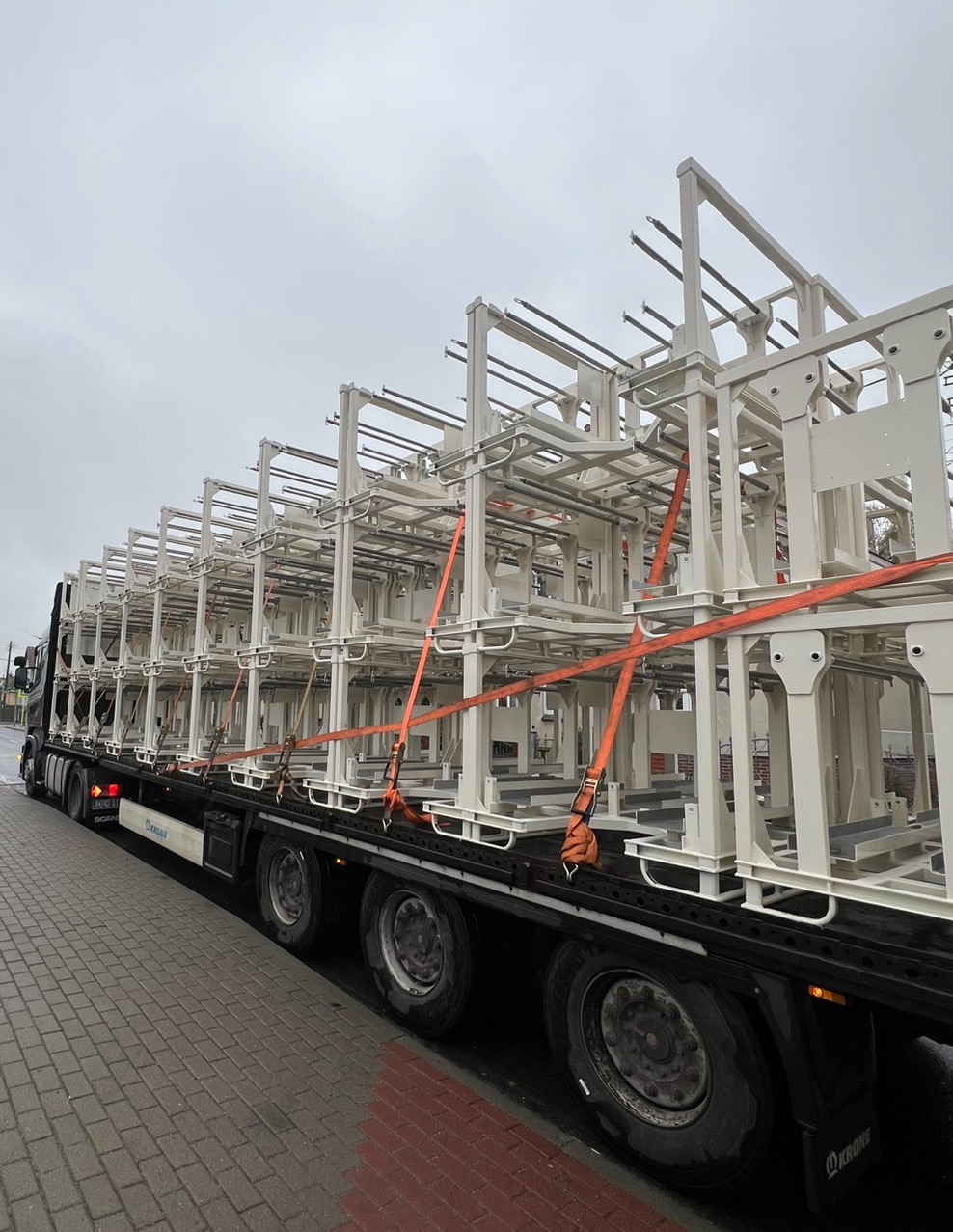 Ciężarówka przewozi metalowe konstrukcje, prawdopodobnie elementy balustrad lub ogrodzeń, zabezpieczone pomarańczowymi pasami. Pochmurne niebo w tle.