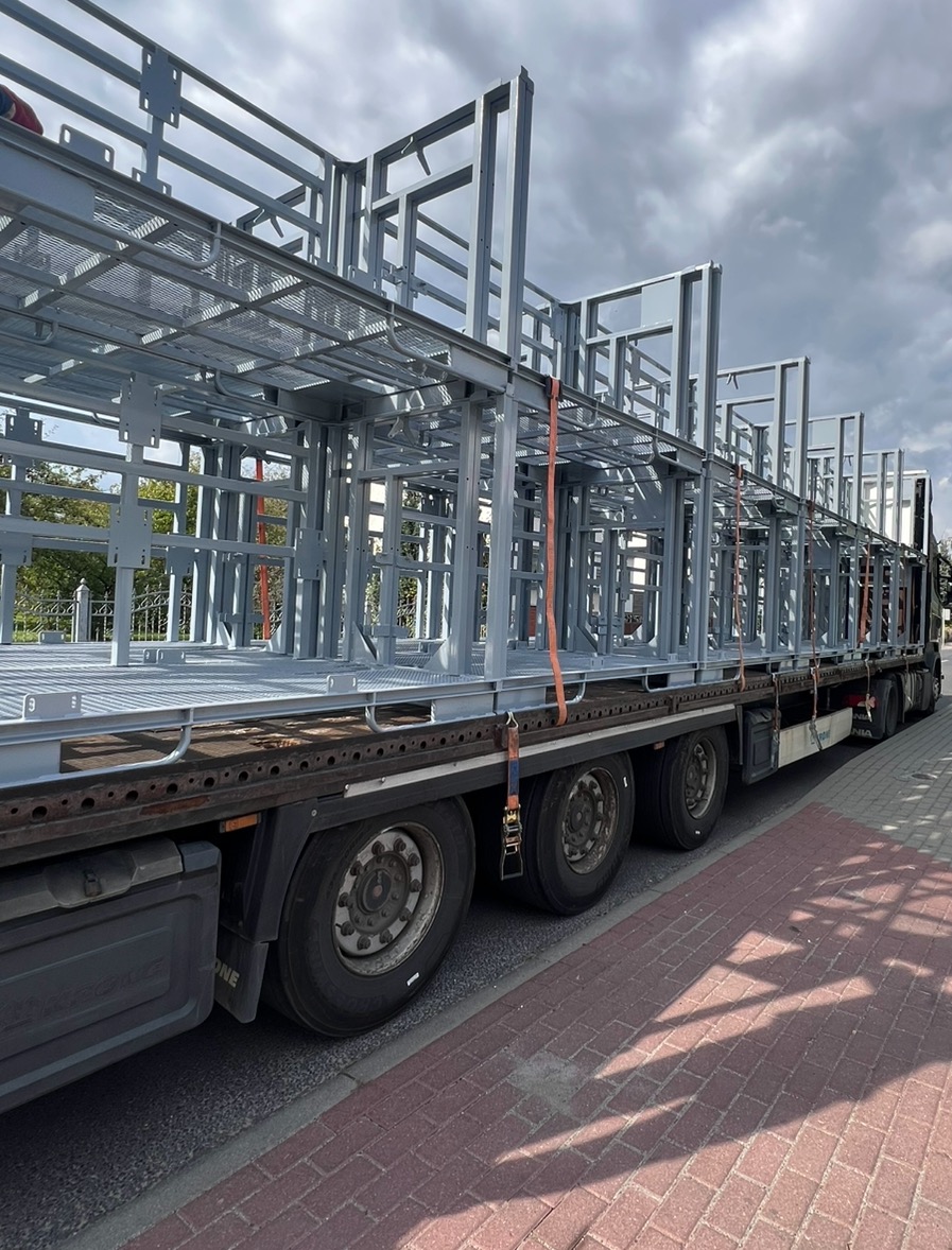 Konstrukcja stalowa, prawdopodobnie balkonowa, zabezpieczona pasami transportowymi na naczepie ciężarówki, gotowa do montażu. Widok z boku, pochmurne niebo w tle.