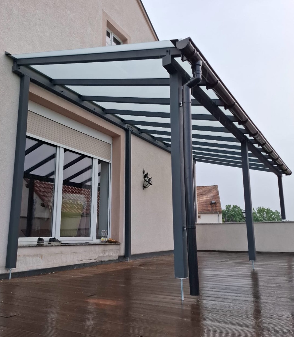 Zadaszenie tarasu z szarą konstrukcją metalową i przezroczystym dachem, widoczne z zewnątrz, na mokrej drewnianej podłodze. Minimalistyczny design.