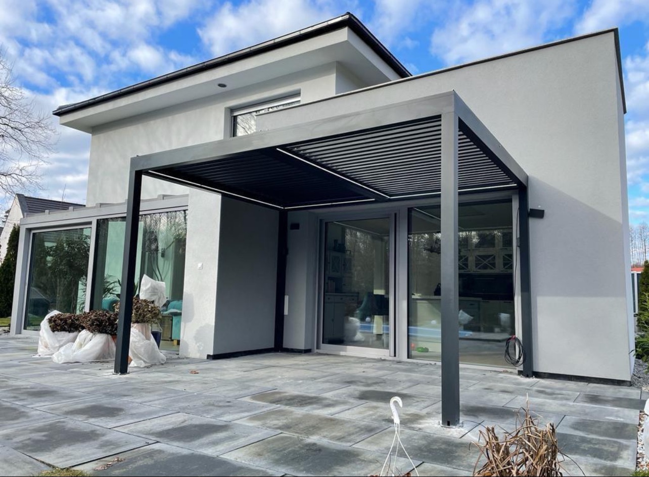 Nowoczesna, minimalistyczna pergola z metalu w kolorze antracytowym, przylegająca do budynku w Żarach. Widoczne szare płyty chodnikowe i fragment ogrodu.