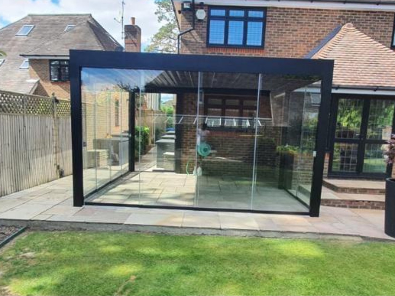 Nowoczesna zabudowa balkonu z czarną ramą i szklanymi ścianami, widok z zewnątrz na tle zadbanego ogrodu i ceglanego domu. Minimalistyczny design, jasne wnętrze.