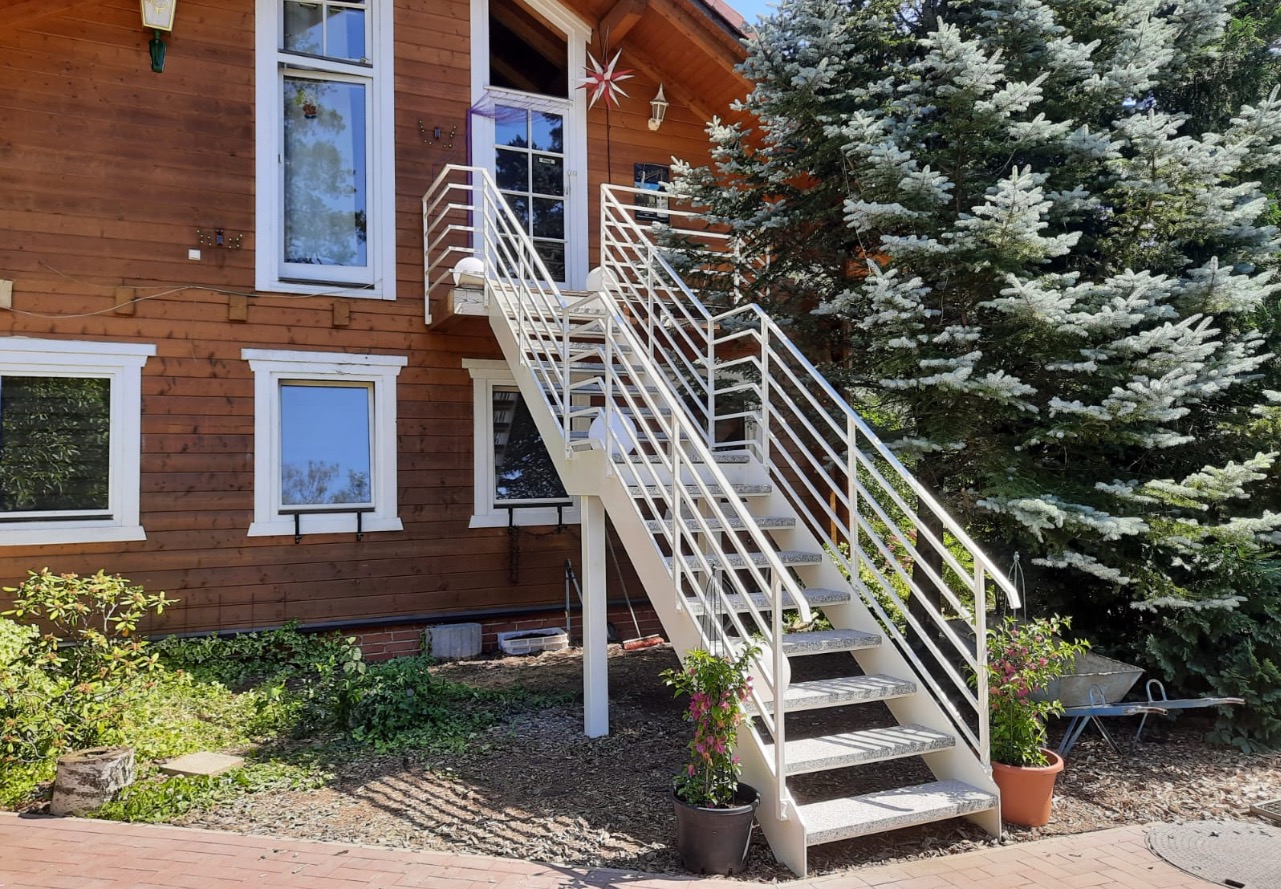 Białe schody zewnętrzne z metalową balustradą prowadzące do drzwi na piętrze drewnianego domu, otoczone zielenią i kwiatami w donicach. Ujęcie z dołu.