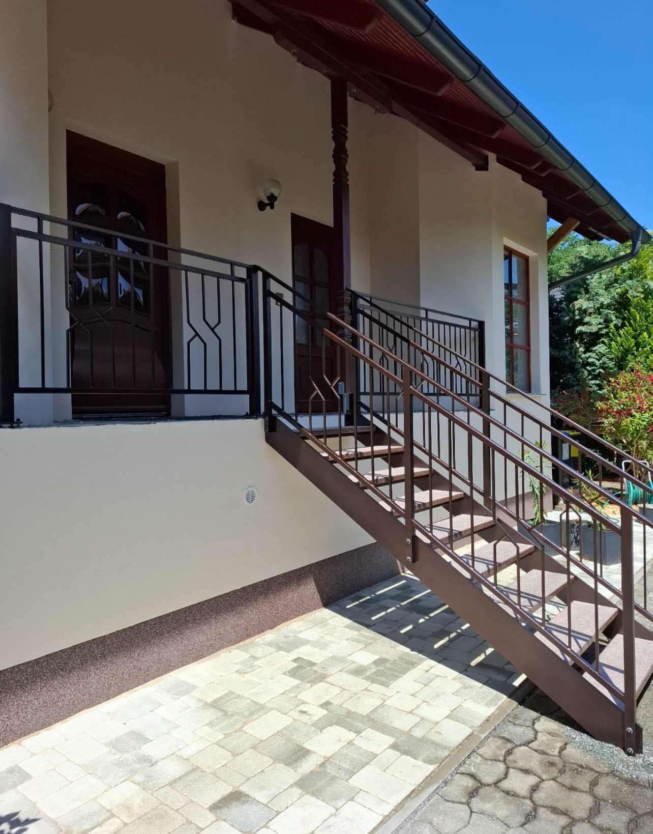 Zewnętrzne schody metalowe z balustradą o geometrycznym wzorze, prowadzące do drzwi wejściowych domu. Brązowa konstrukcja na tle jasnej elewacji i kostki brukowej.