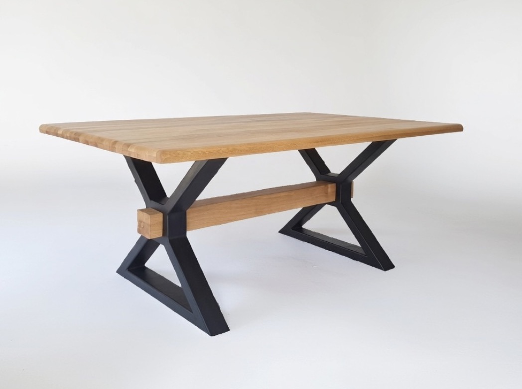 Stół z blatem z jasnego drewna i czarnymi, metalowymi nogami w kształcie litery X, połączonymi drewnianą belką. Minimalistyczny design na białym tle.