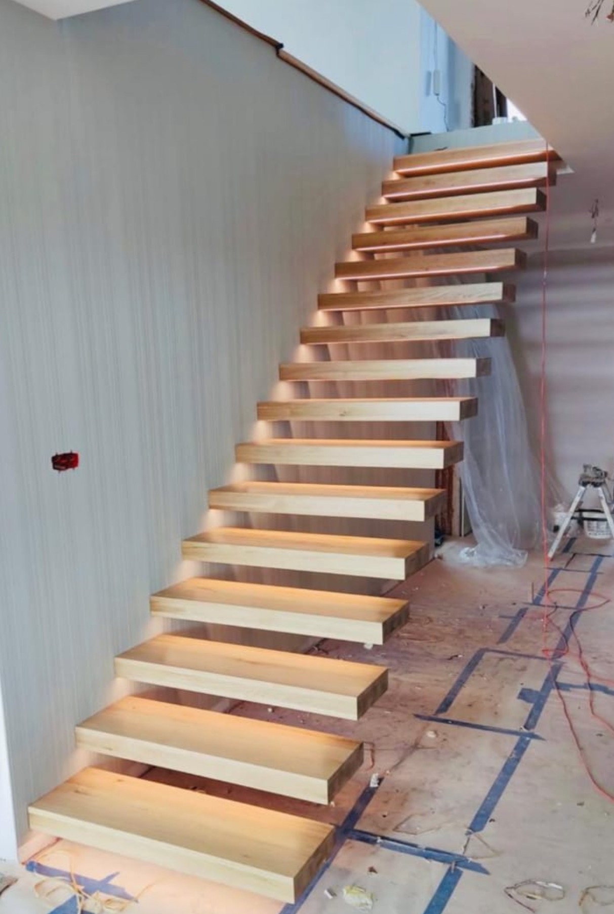 Drewniane schody wiszące z podświetleniem LED, zamontowane przy szarej ścianie. Widoczne przewody i narzędzia sugerują etap wykończeniowy.