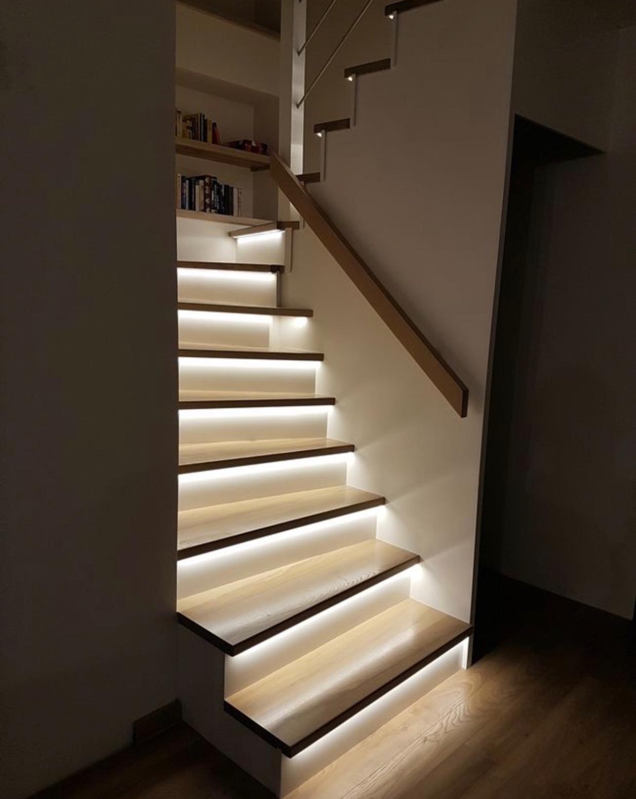 Drewniane schody z podświetleniem LED, widok z dołu. Jasne stopnie z ciemnymi krawędziami, drewniana poręcz, półka z książkami w tle. Nowoczesny design, ciepłe oświetlenie.