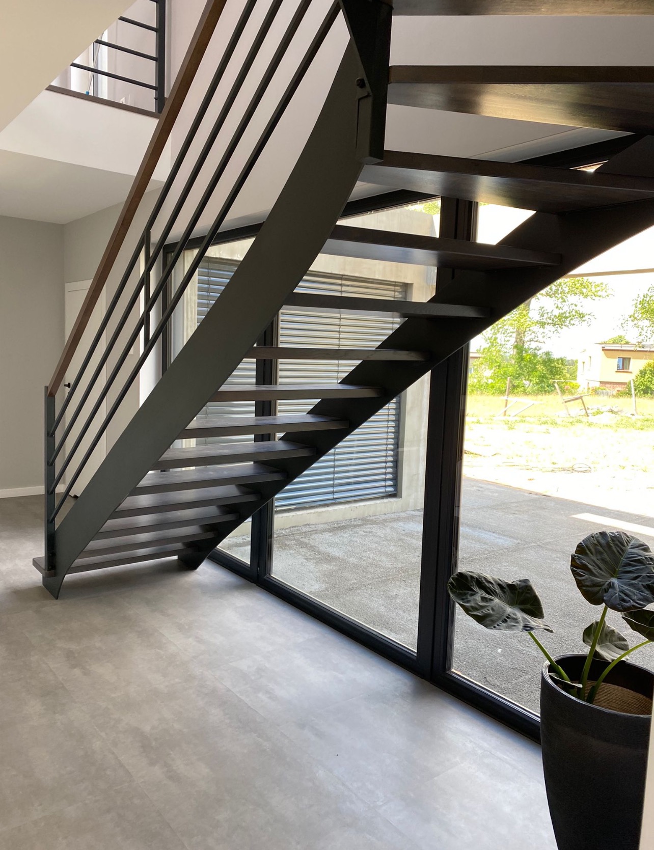 Nowoczesne schody metalowe z drewnianymi stopniami, minimalistyczna balustrada, duże okno z widokiem na zieleń, donica z rośliną w rogu. Jasne wnętrze, szara podłoga.