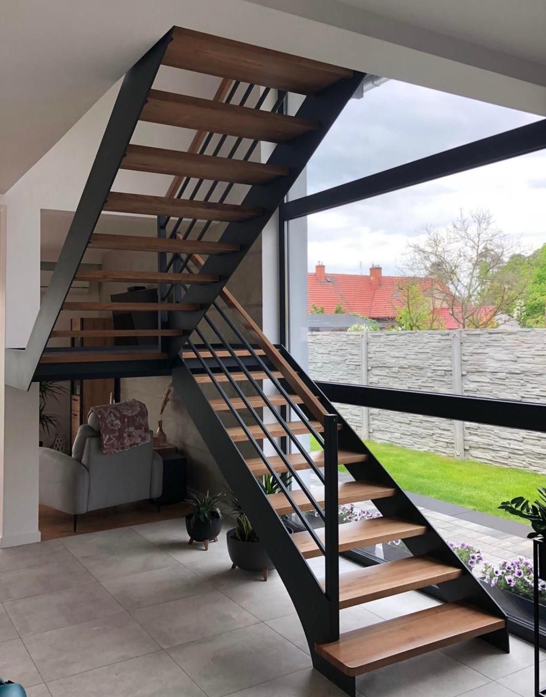 Nowoczesne schody metalowe z drewnianymi stopniami w jasnym wnętrzu z dużym oknem. Minimalistyczny design, czarna konstrukcja, widok na ogród.