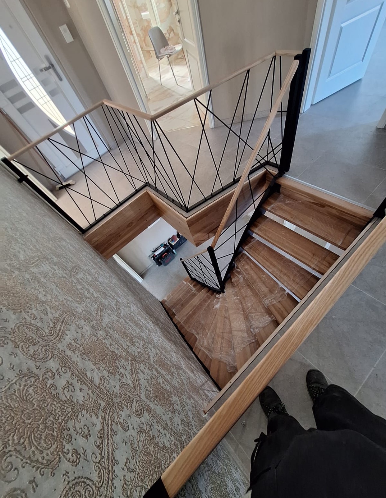 Spiralne schody z drewnianymi stopniami i nowoczesną balustradą z czarnych, krzyżujących się prętów, widziane z góry, z fragmentem osoby w kadrze.