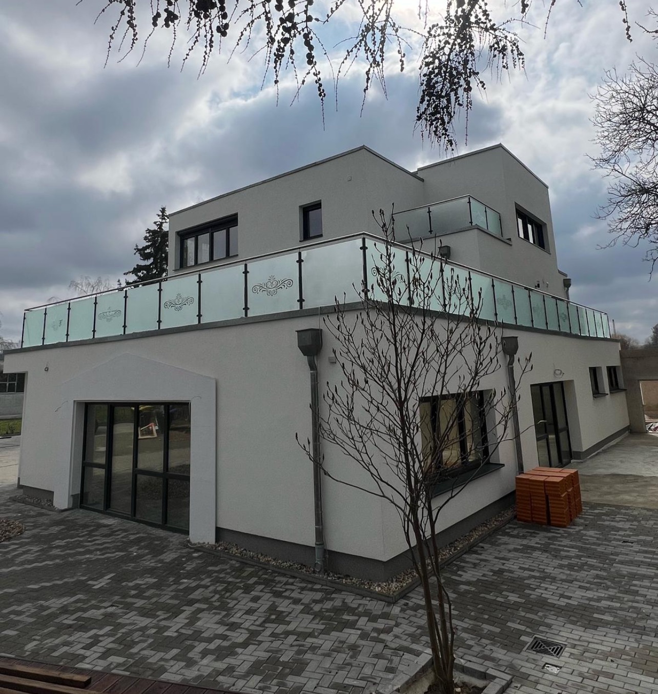 Nowoczesny dom z balkonem zabezpieczonym szklaną balustradą z delikatnym wzorem, widok z poziomu brukowanego podjazdu, pochmurne niebo w tle.