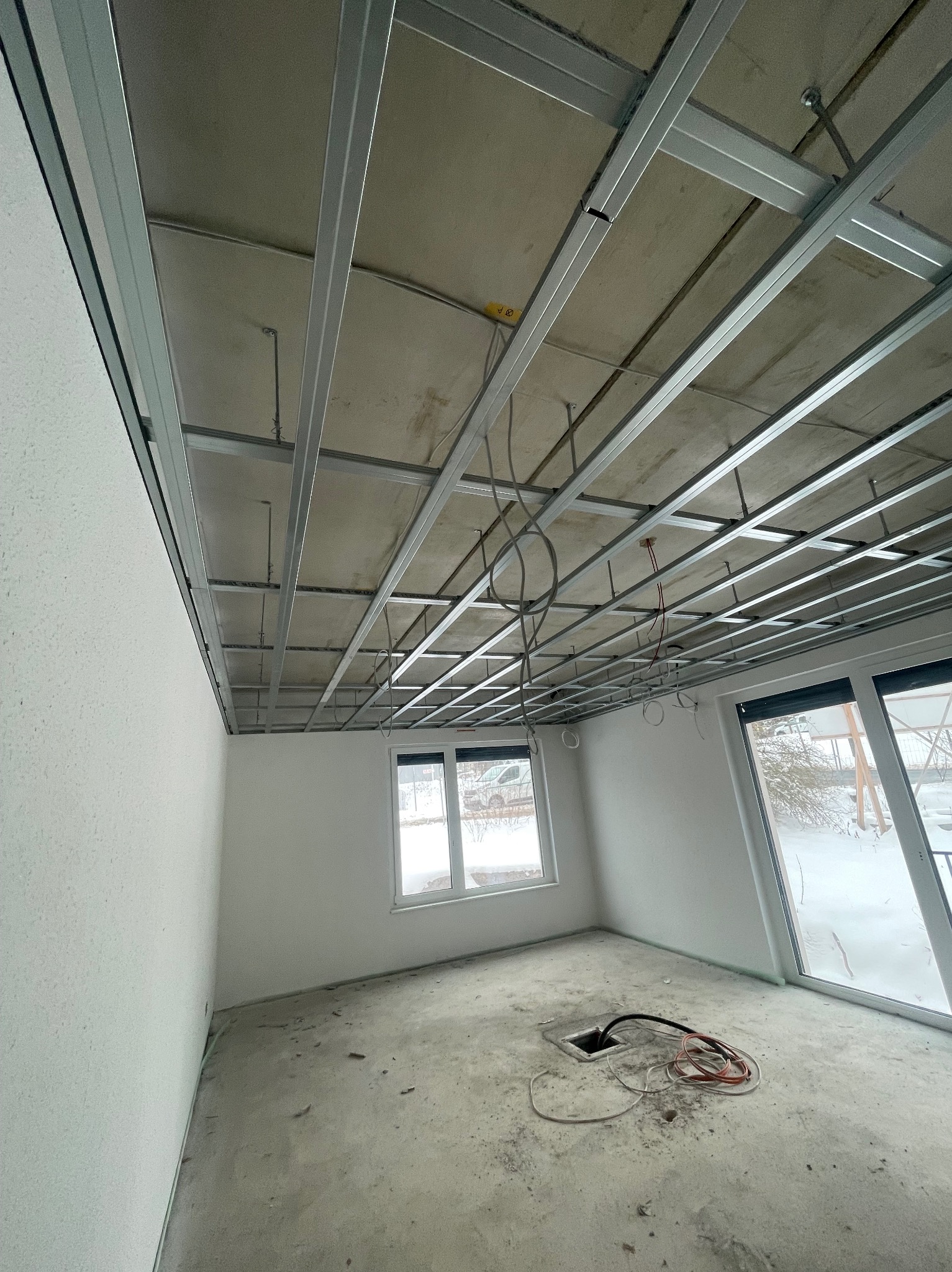 Sufit podwieszany z metalową konstrukcją w surowym wnętrzu. Widoczne okno i przewody. Etap prac wykończeniowych w pomieszczeniu z oknami i betonową podłogą.