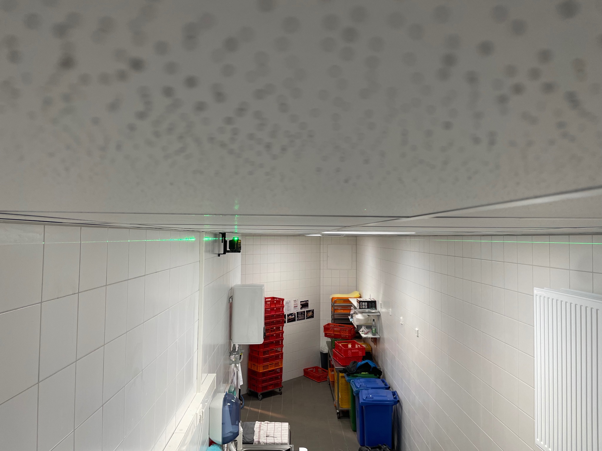 Białe płytki w pomieszczeniu z podwieszanym sufitem, poziomica laserowa wyznacza linię. Widoczne pojemniki i regały.