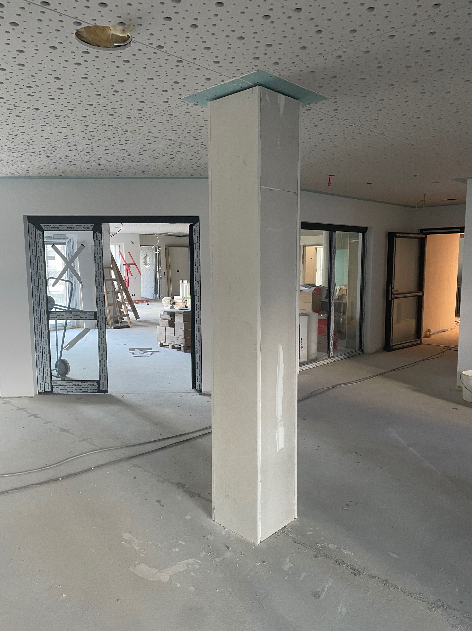Wnętrze w trakcie prac wykończeniowych: kolumna obłożona płytami gipsowo-kartonowymi, widoczne otwarte drzwi i sufit z perforacją. Stan surowy, przygotowanie do dalszych prac.