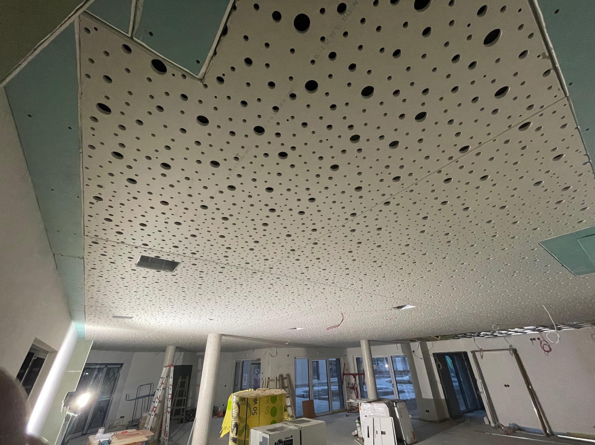 Sufit z płyty akustycznej z perforacją w nowym biurze, widok z dołu. Widoczne elementy konstrukcyjne i przygotowania do dalszych prac wykończeniowych.