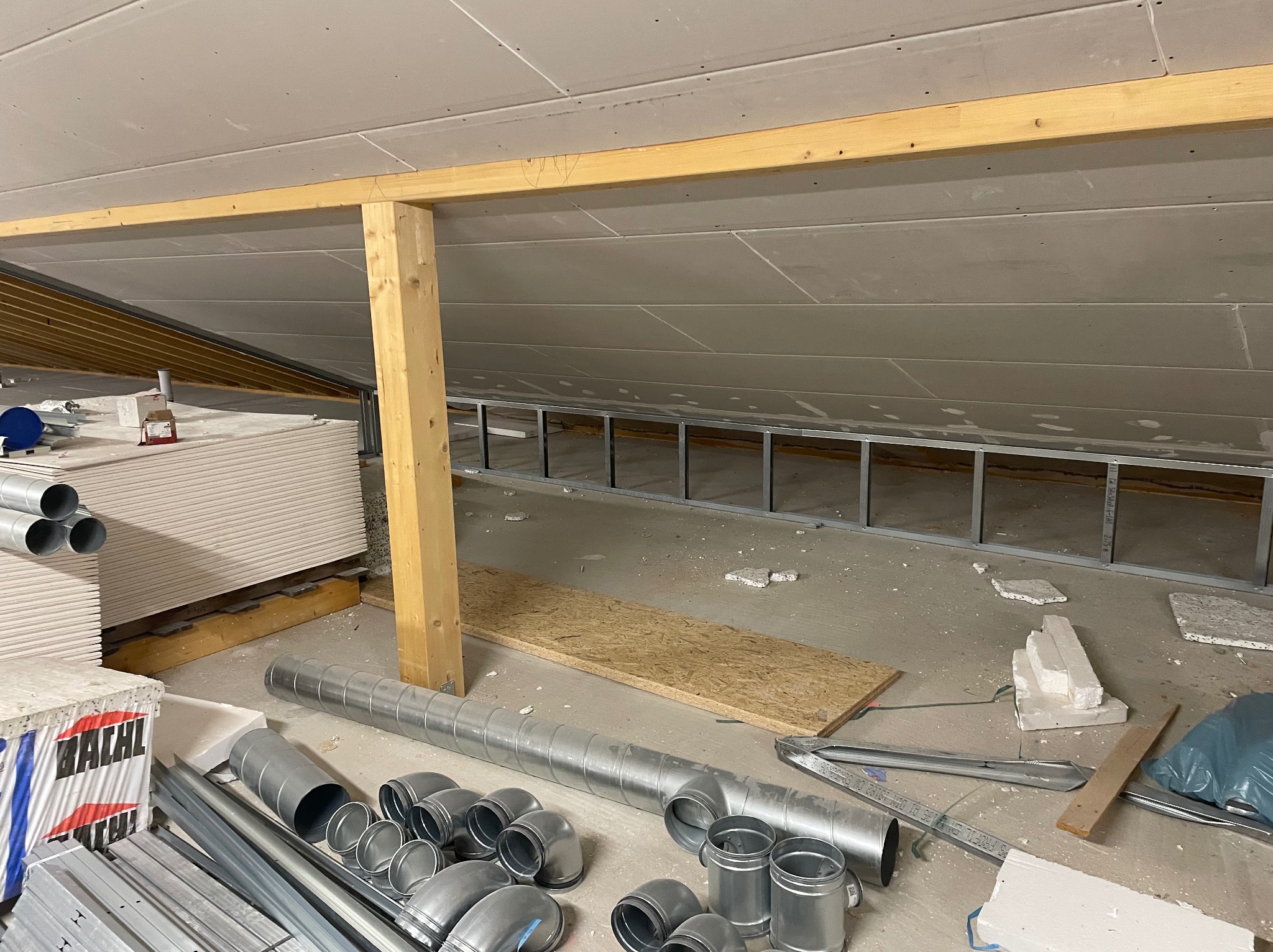 Poddasze w trakcie adaptacji: widoczne płyty karton-gips na skośnym suficie, metalowa konstrukcja, drewniane belki i elementy wentylacji. Etap prac wykończeniowych.