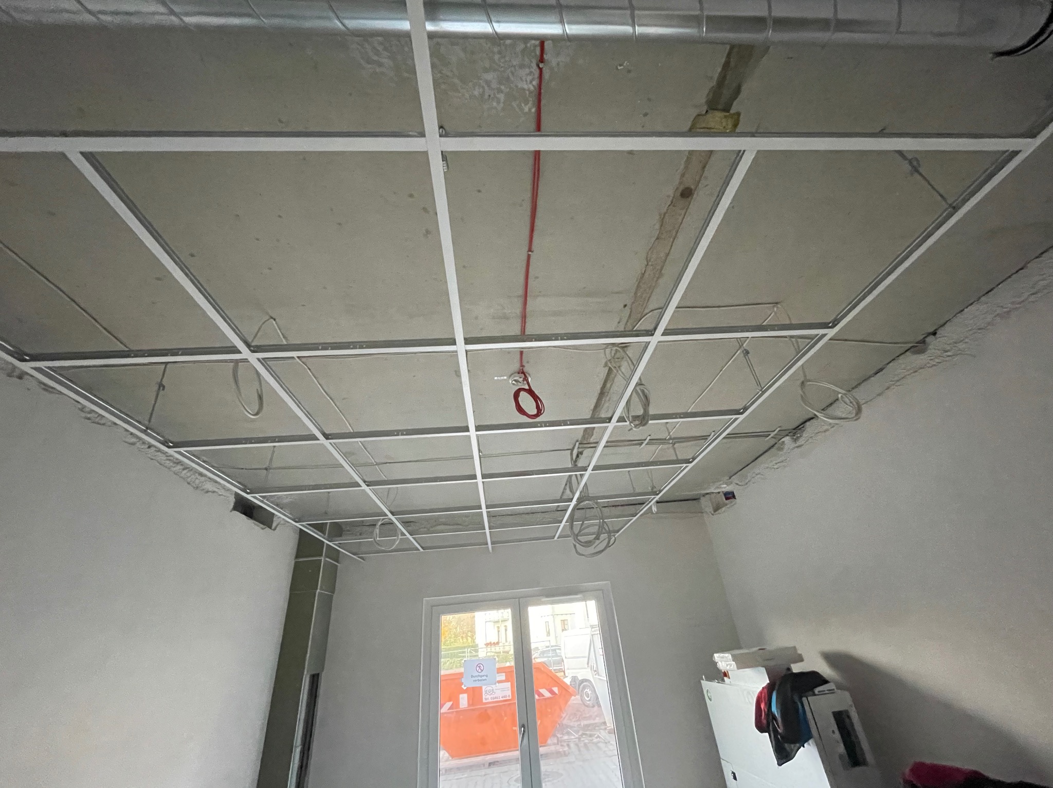 Montaż stelaża sufitu podwieszanego z widocznymi przewodami elektrycznymi i kanałem wentylacyjnym. W tle okno z kontenerem na gruz.