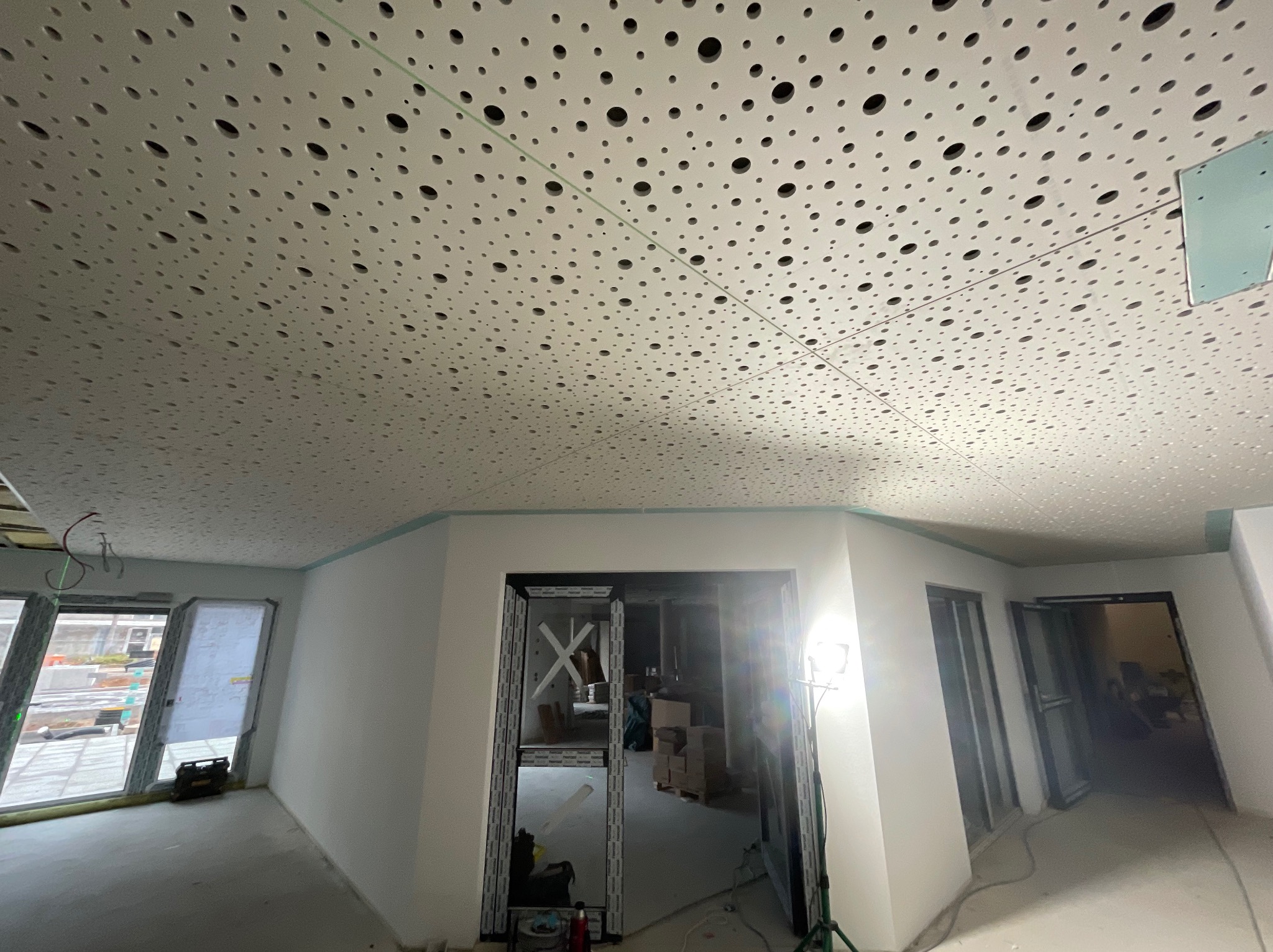 Sufit z perforowanej płyty gipsowej w trakcie montażu, widoczne profile i przewody. W tle surowe ściany i otwory drzwiowe. Oświetlenie budowlane.