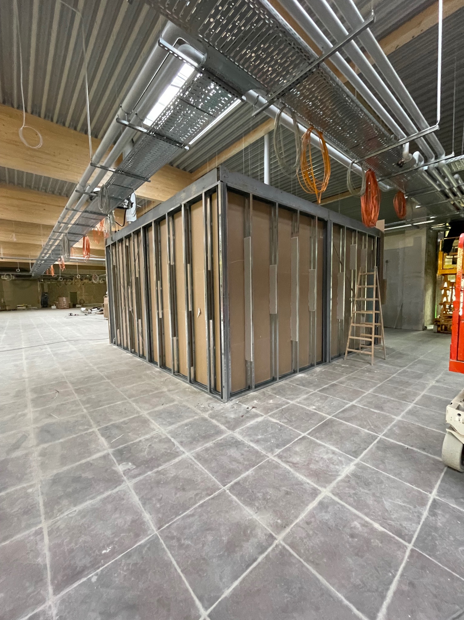 Wnętrze w trakcie budowy ścian działowych z płyt kartonowo-gipsowych na metalowym stelażu. Widoczne instalacje pod sufitem i drabina. Surowy stan wykończenia podłogi.