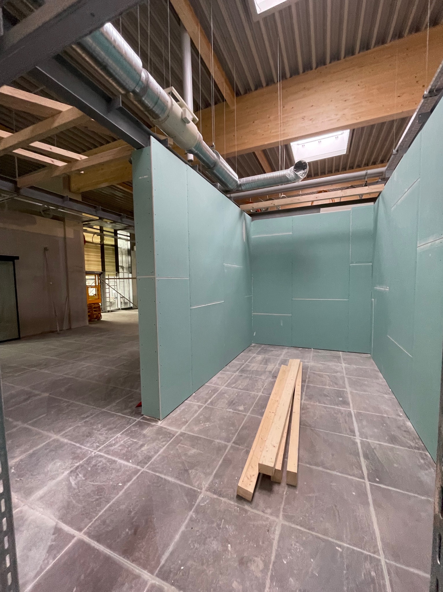 Montaż ścian działowych z płyt gipsowo-kartonowych w surowym wnętrzu. Widoczne drewniane belki stropowe i wentylacja. Na podłodze leżą drewniane kantówki.