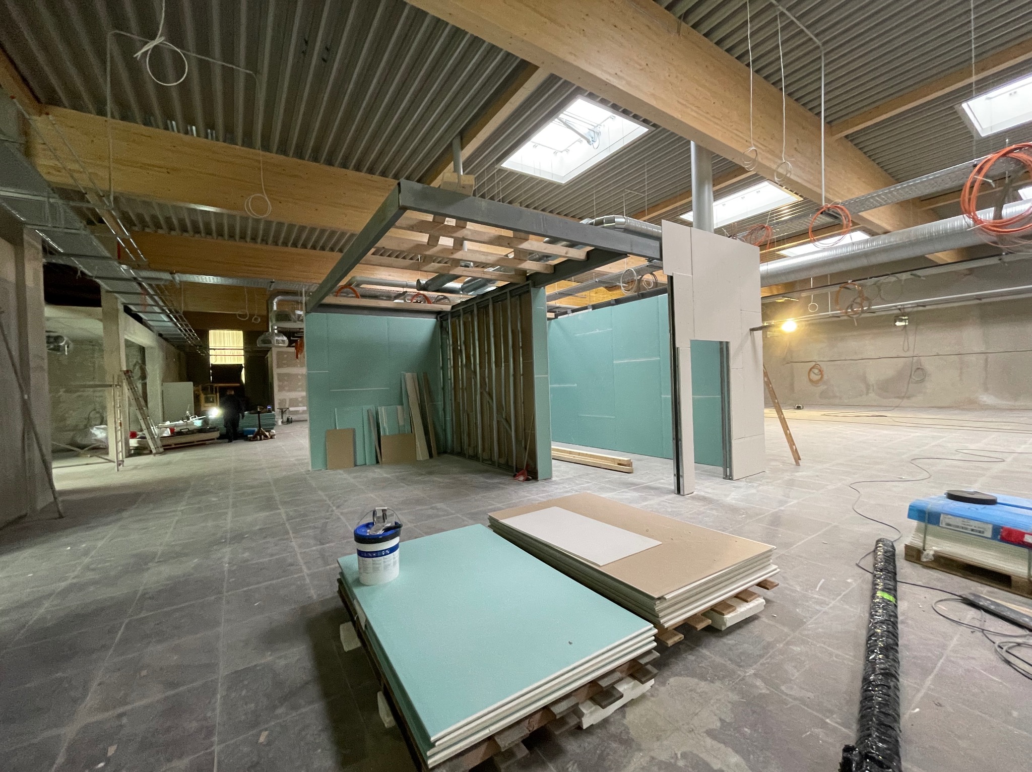 Wnętrze w trakcie prac z płytami gipsowo-kartonowymi, widoczne stelaże i przygotowane płyty. Oświetlenie sufitowe i instalacje pod stropem. Surowy stan wykończenia.