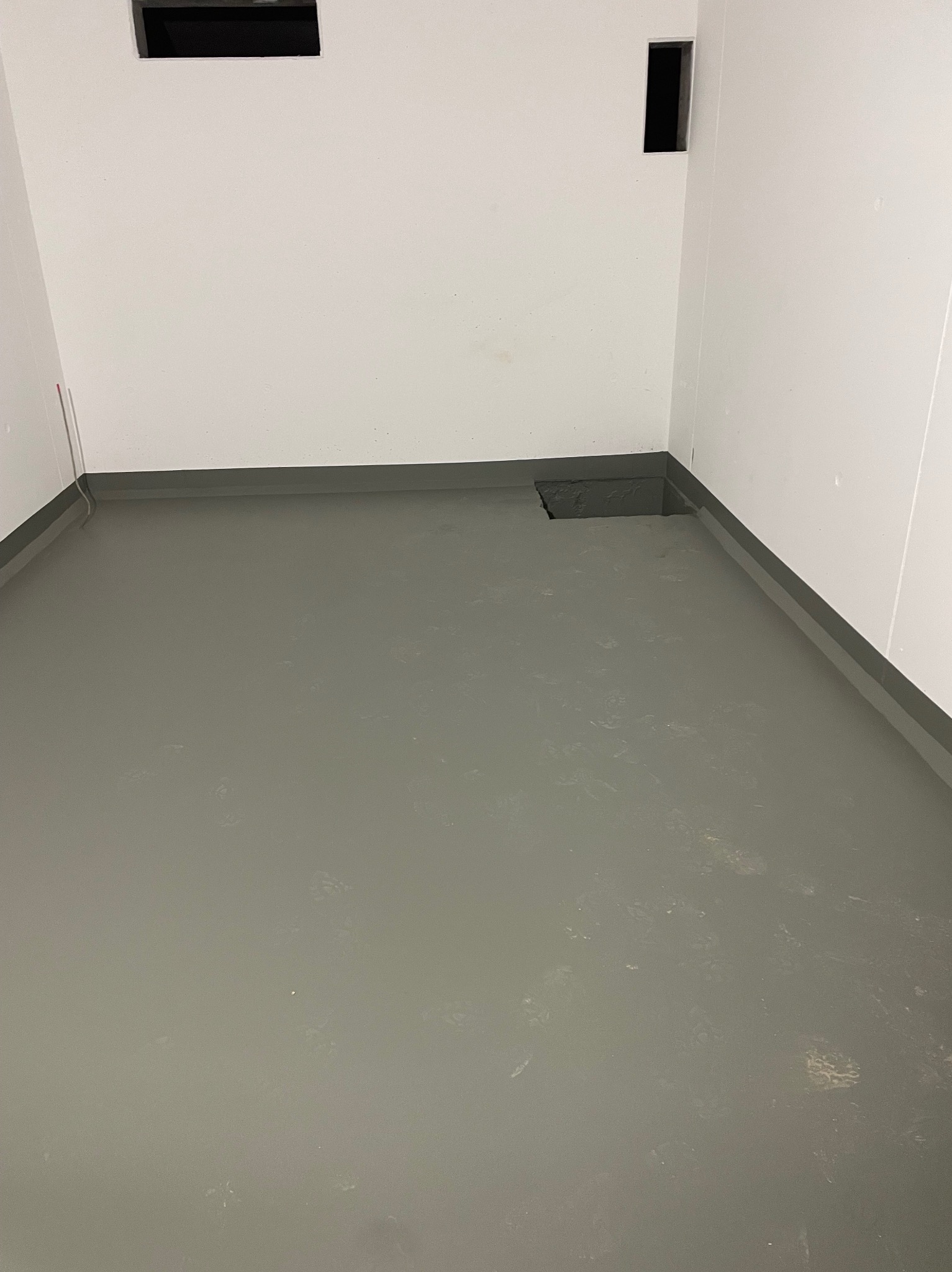 Szare, jednolite wykończenie podłogi w pomieszczeniu z białymi ścianami i dwoma otworami wentylacyjnymi. Widoczne ślady po pracy na podłodze.