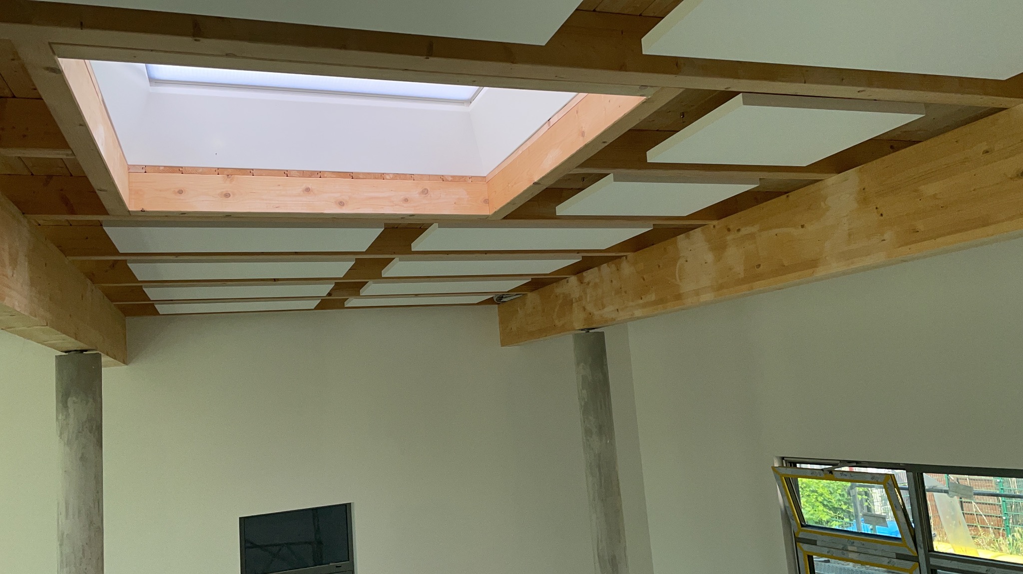Drewniany strop z oknem dachowym i białymi panelami akustycznymi. Widok z dołu, surowy betonowy filar, okno w tle. Jasne wnętrze w trakcie wykończenia.