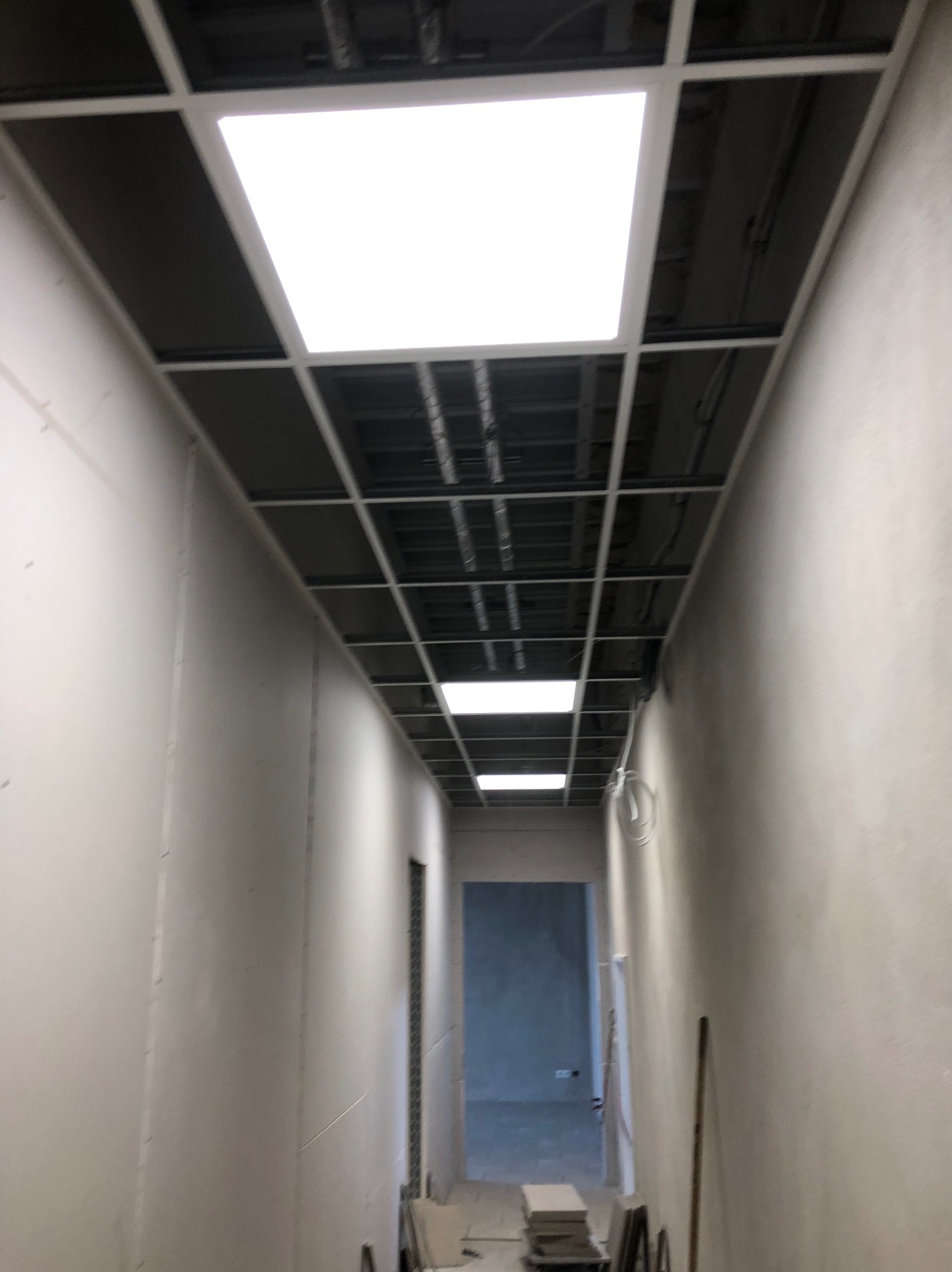 Korytarz w trakcie wykańczania: sufit podwieszany z oświetleniem rastrowym, ściany przygotowane do malowania, widoczne narzędzia. Perspektywa z dołu.