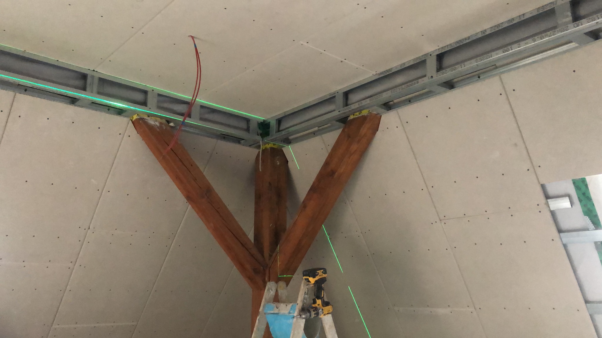 Sufit z płyt gipsowo-kartonowych w trakcie montażu, widoczne drewniane belki, metalowy stelaż i poziomica laserowa. W tle drabina i wkrętarka.