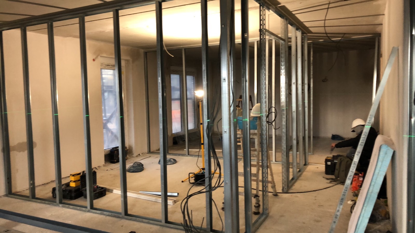 Metalowa konstrukcja ścian działowych w surowym wnętrzu, widoczne narzędzia i pracownik w kasku. Laserowe poziomice wyznaczają linie. Etap budowy ścian z płyt g-k.