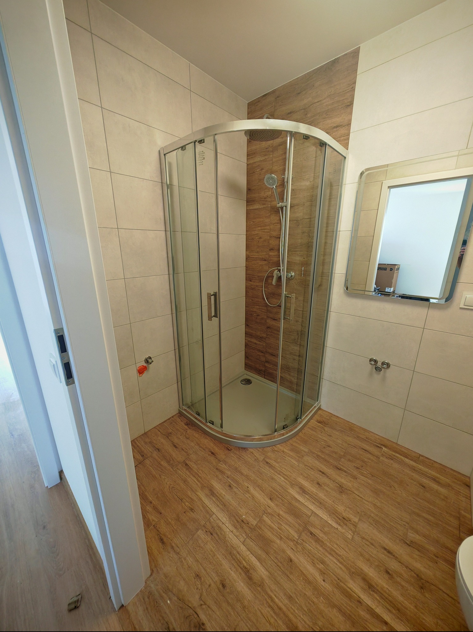 Nowoczesna łazienka po remoncie: kabina prysznicowa z drewnopodobnymi płytkami, lustro z oświetleniem i podłoga imitująca drewno. Widok z perspektywy drzwi.