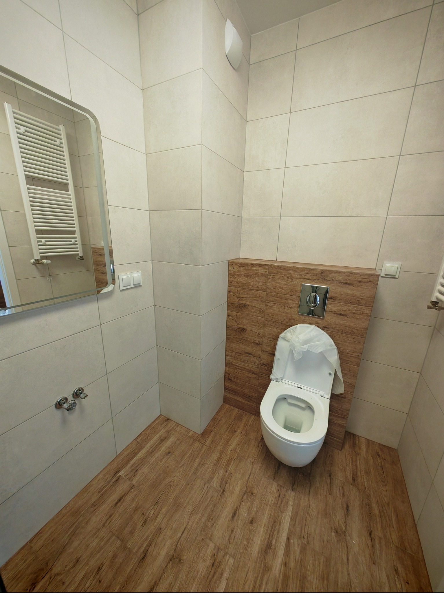 Nowoczesna łazienka po remoncie: beżowe płytki ścienne, drewnopodobna podłoga, wisząca toaleta z drewnianym panelem, lustro z grzejnikiem. Widok z perspektywy.