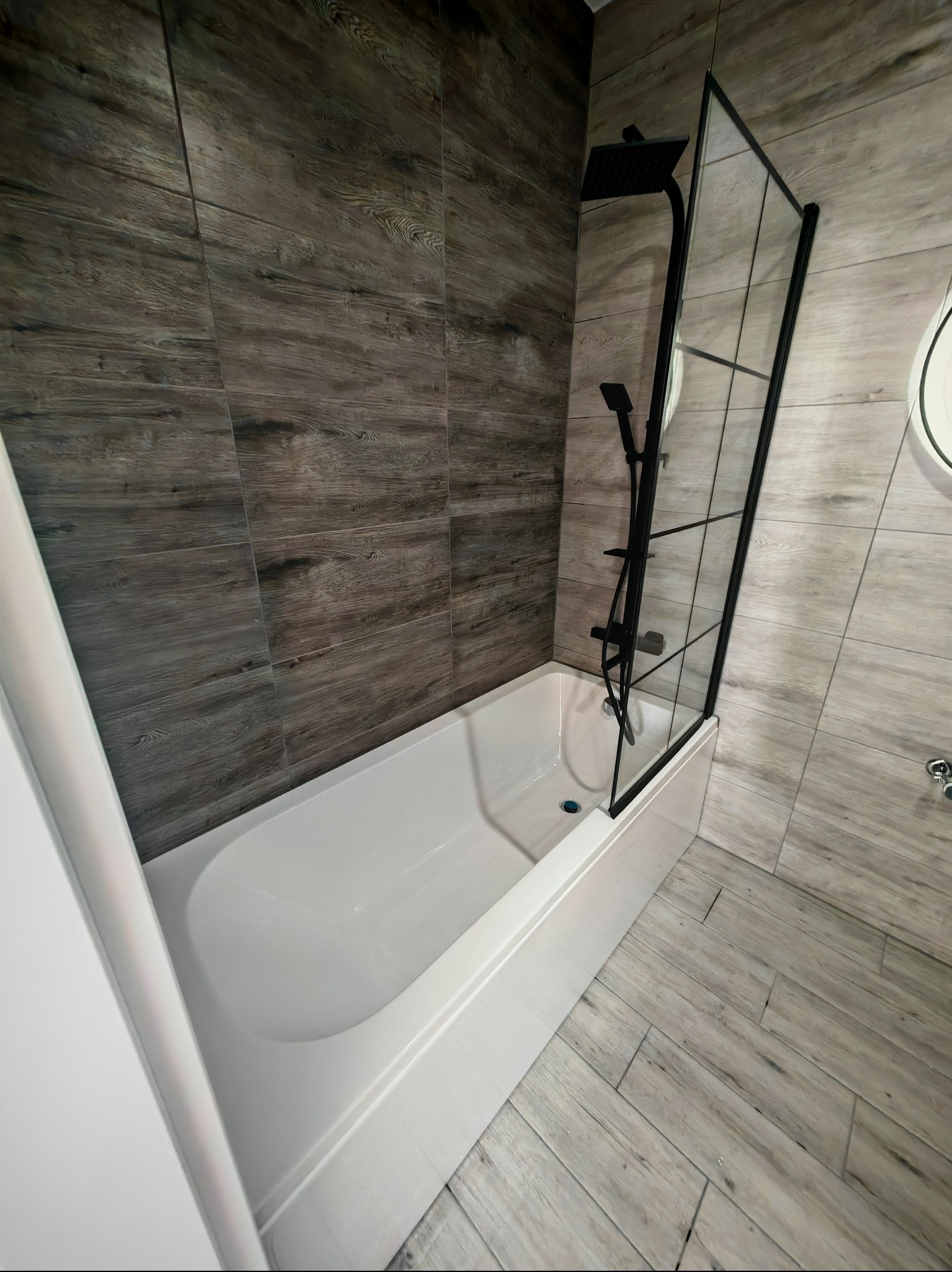 Nowoczesna łazienka z wanną, czarną armaturą i szklanym parawanem. Płytki imitujące drewno na ścianach i podłodze. Minimalistyczny design, widok z góry.