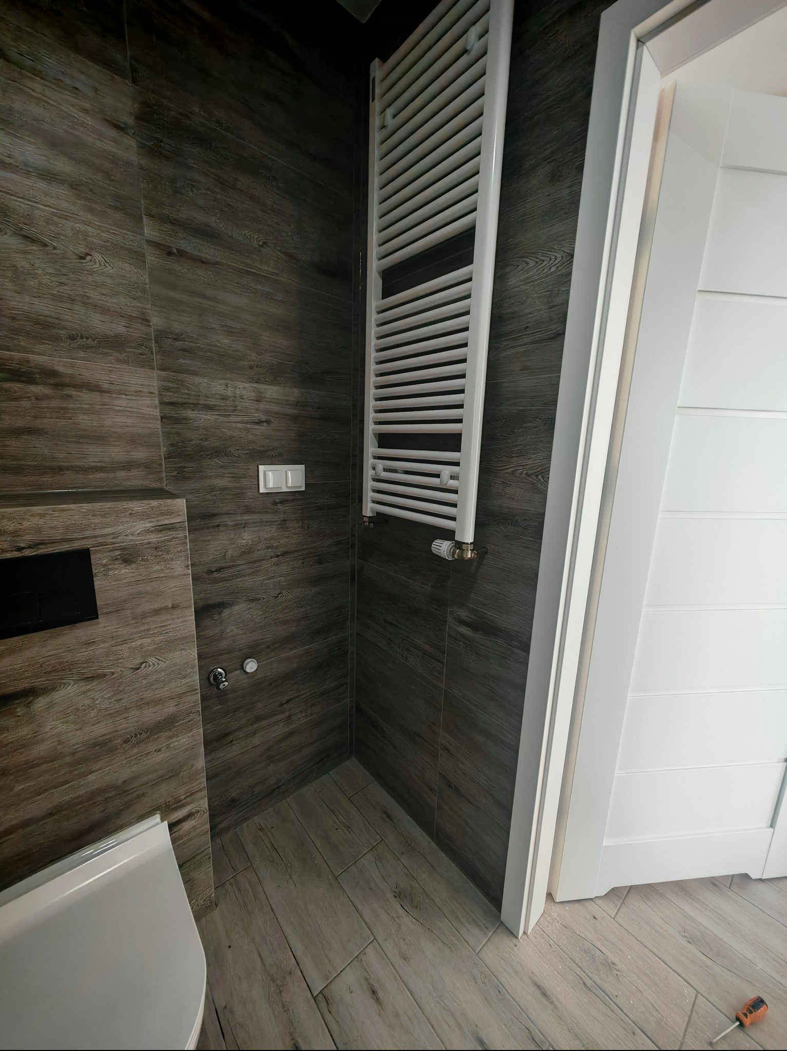 Wykończona łazienka z brązowymi płytkami imitującymi drewno, białą drabinką grzejnikową i częściowo widoczną toaletą. Widoczny fragment białych drzwi.