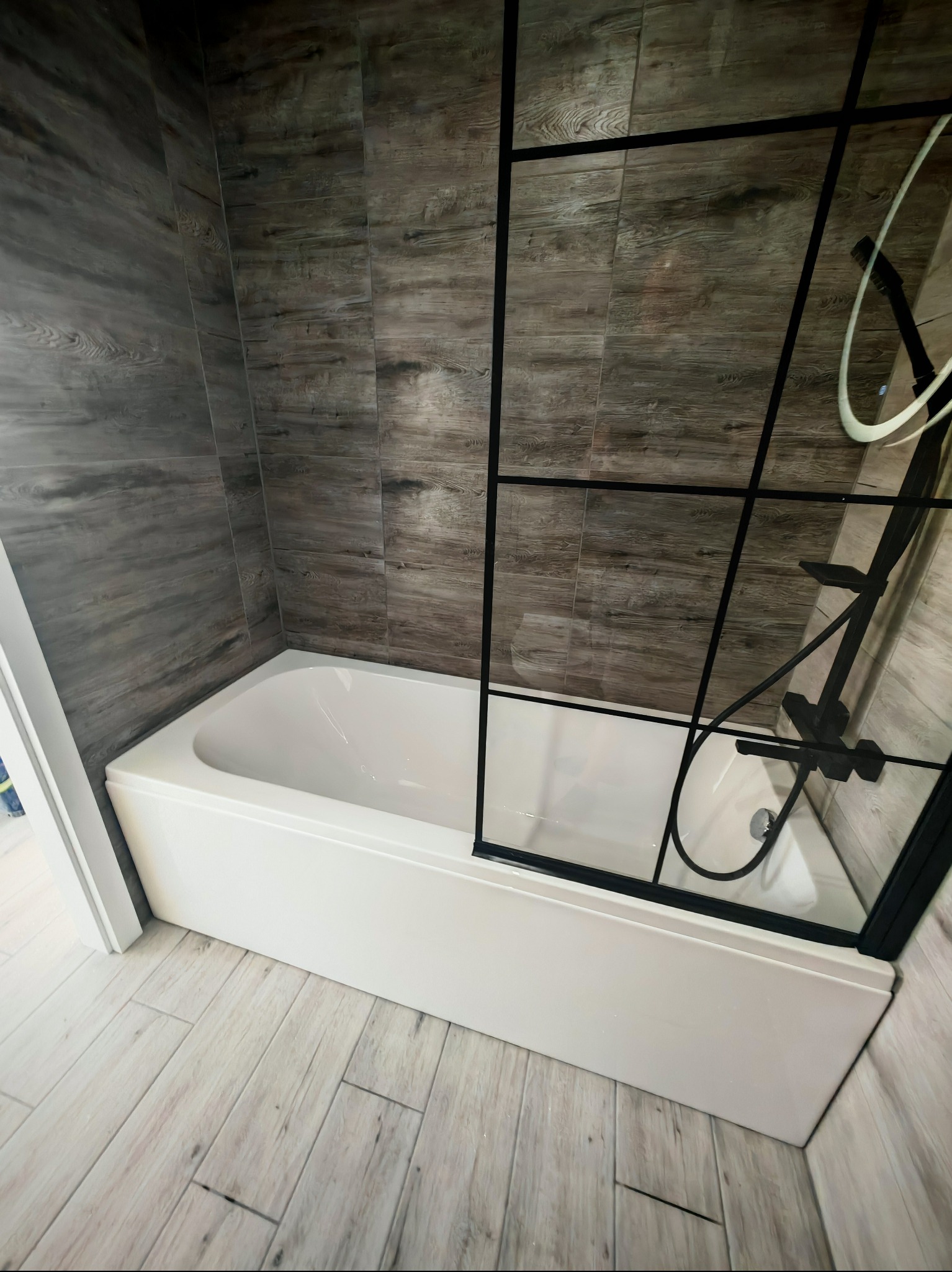 Nowoczesna łazienka z wanną i kabiną prysznicową w stylu loft, wykończona płytkami imitującymi drewno. Czarna armatura i szklana ścianka kabiny.