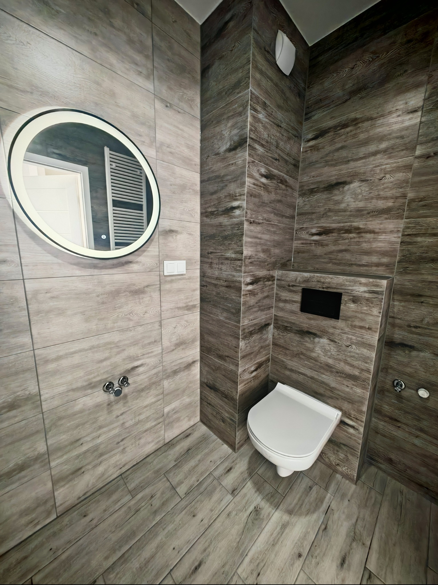 Nowoczesna łazienka z drewnopodobnymi płytkami, okrągłym lustrem z podświetleniem LED i podwieszaną toaletą. Widok na fragment wnętrza z perspektywy podłogi.