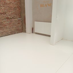 DZDekor - Biała posadzka żywiczna w surowym wnętrzu z ceglaną ścianą i napisem 'Palais Blanc' na ścianie. Widoczne rury grzewcze. Minimalistyczny design.