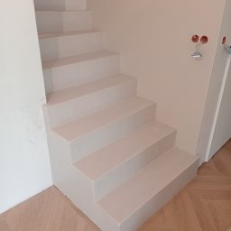 DZDekor - Wykończone, minimalistyczne schody w jasnym odcieniu, z widocznymi gniazdkami elektrycznymi na ścianie i parkietem u dołu. Ujęcie z dołu, podkreślające geometrię stopni.