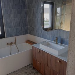 DZDekor - Remont łazienki - Nowoczesna łazienka z niebieskimi płytkami, białą wanną i umywalką na drewnianej szafce z ozdobnymi uchwytami. Lustro o falistym kształcie odbija okno.