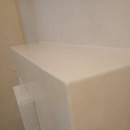 DZDekor - Remont łazienki - Gładka, beżowa półka ścienna z ostrymi krawędziami, wkomponowana w ścianę z jasnych płytek. Widoczny fragment białego, żebrowanego elementu instalacji.