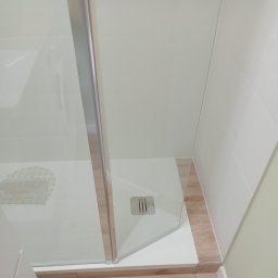 DZDekor - Remont łazienki - Nowoczesna kabina prysznicowa z białym brodzikiem, minimalistycznym odpływem i szklanymi drzwiami, otoczona białymi płytkami i pasem drewnopodobnych płytek. W środku widoczna biała mata...