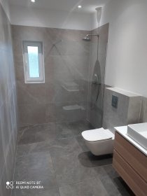 Nowoczesna łazienka z szarymi płytkami imitującymi marmur, kabiną typu walk-in i podwieszaną toaletą. Umywalka z szafką z drewnianym frontem. Minimalistyczne oświetlenie.