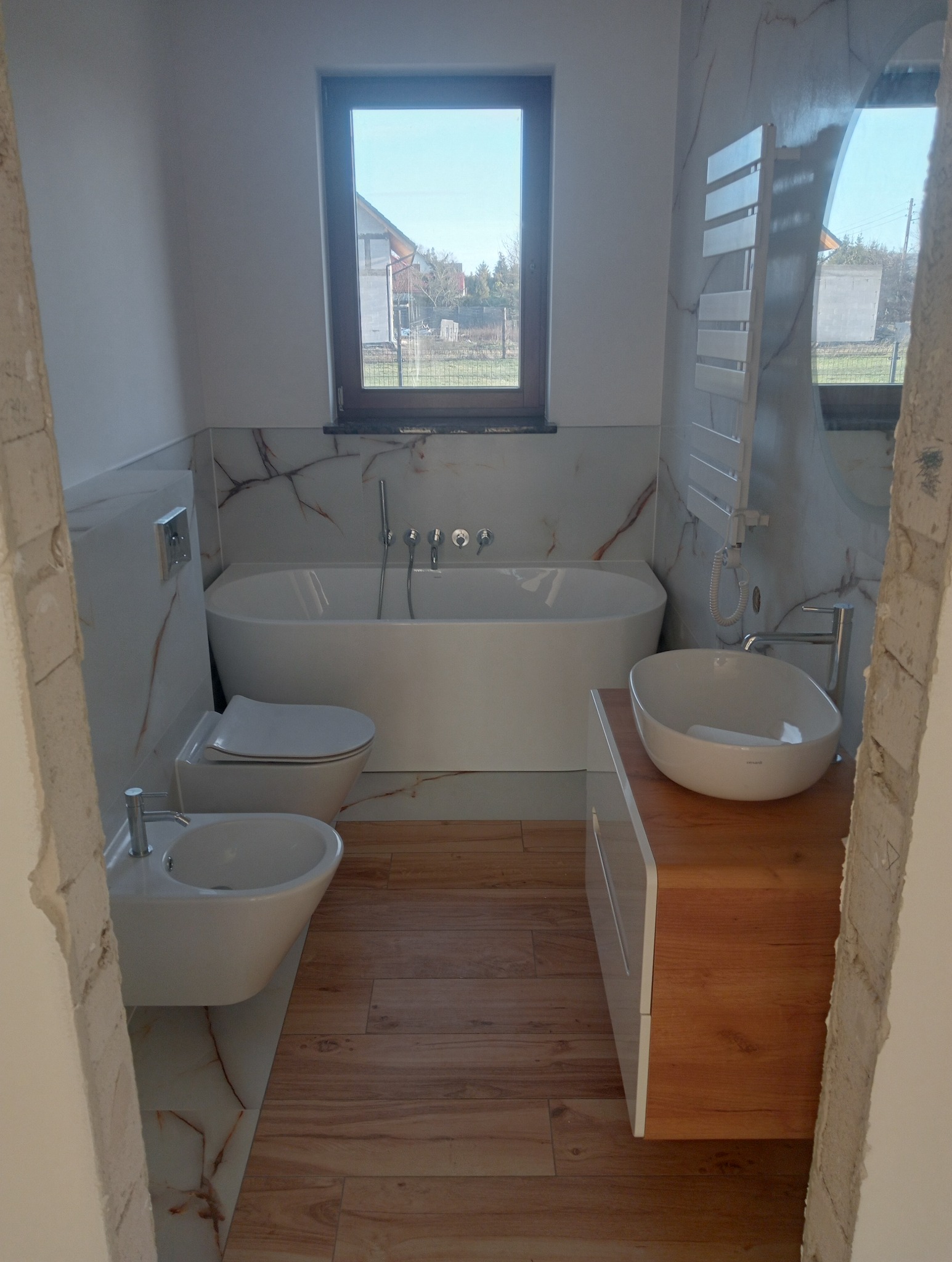 Nowoczesna aranżacja łazienki w Szczecinie: biała wanna, umywalka na drewnianej szafce, bidet i toaleta. Płytki imitujące marmur i drewno tworzą elegancki wystrój.