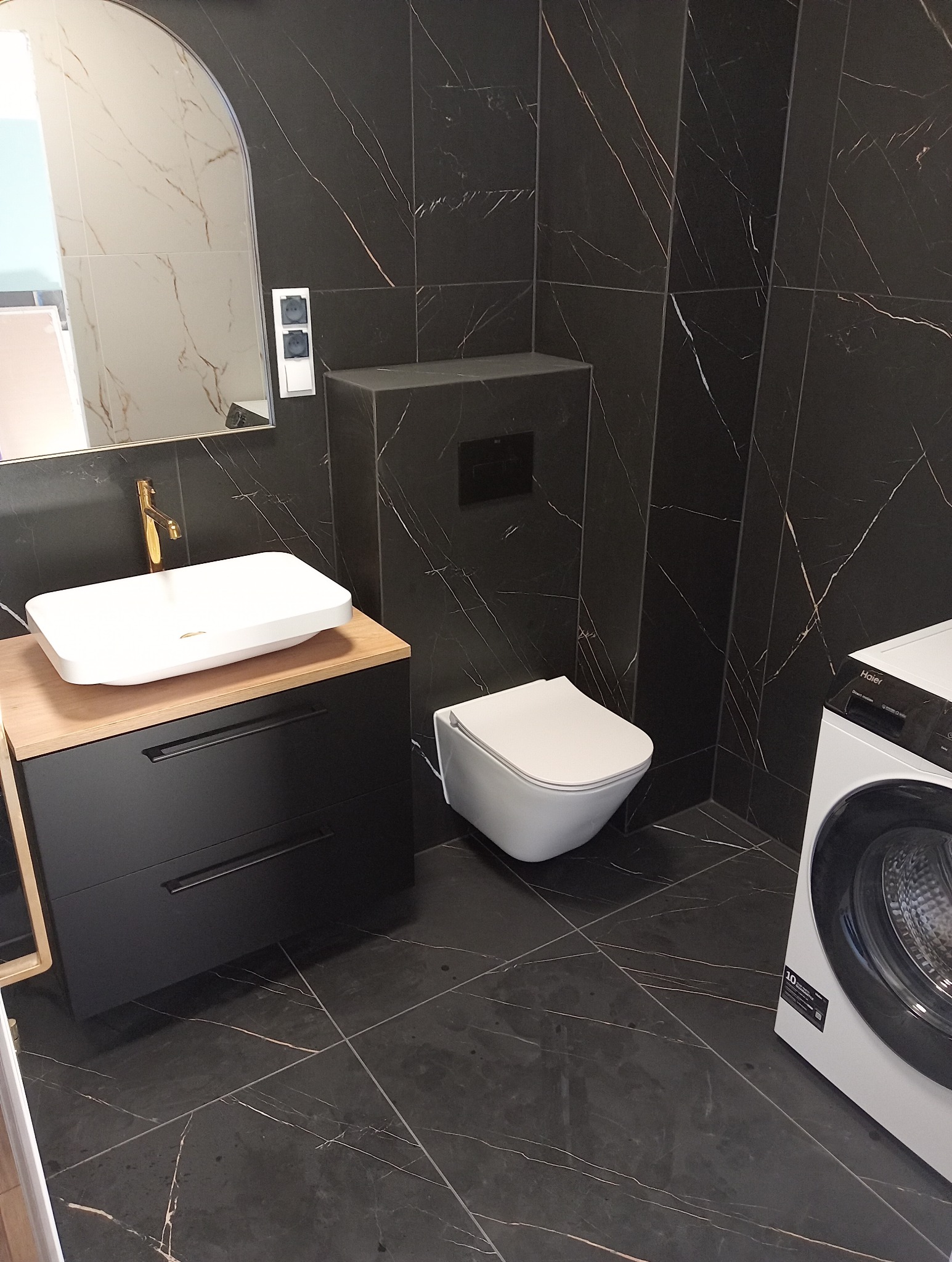 Nowoczesna łazienka z czarnymi płytkami imitującymi marmur, białą umywalką nablatową z złotą armaturą i podwieszaną toaletą. Widoczna pralka.
